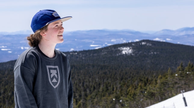 Person wearing a blue cap and dark hoodie with a Killington logo, standing on a mountain with a scenic view.