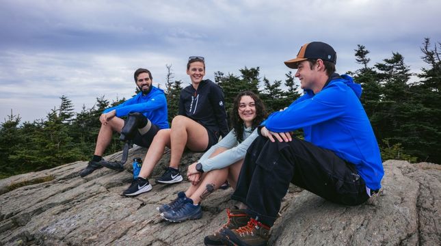 Four people sitting on a rocky outcrop with trees and a cloudy sky in the background