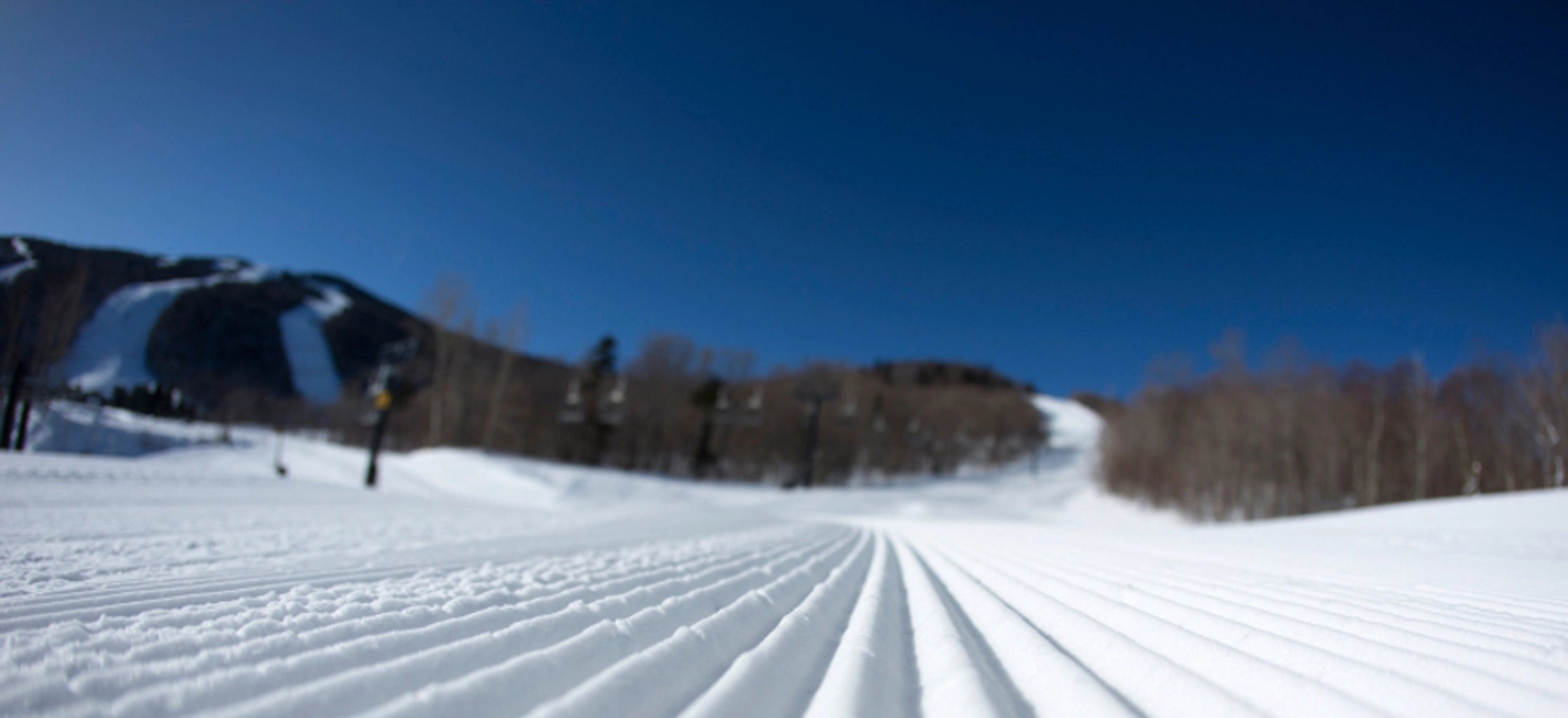 A snowy ski slope with groomed tracks leading toward mountains and bare trees under a clear blue sky.