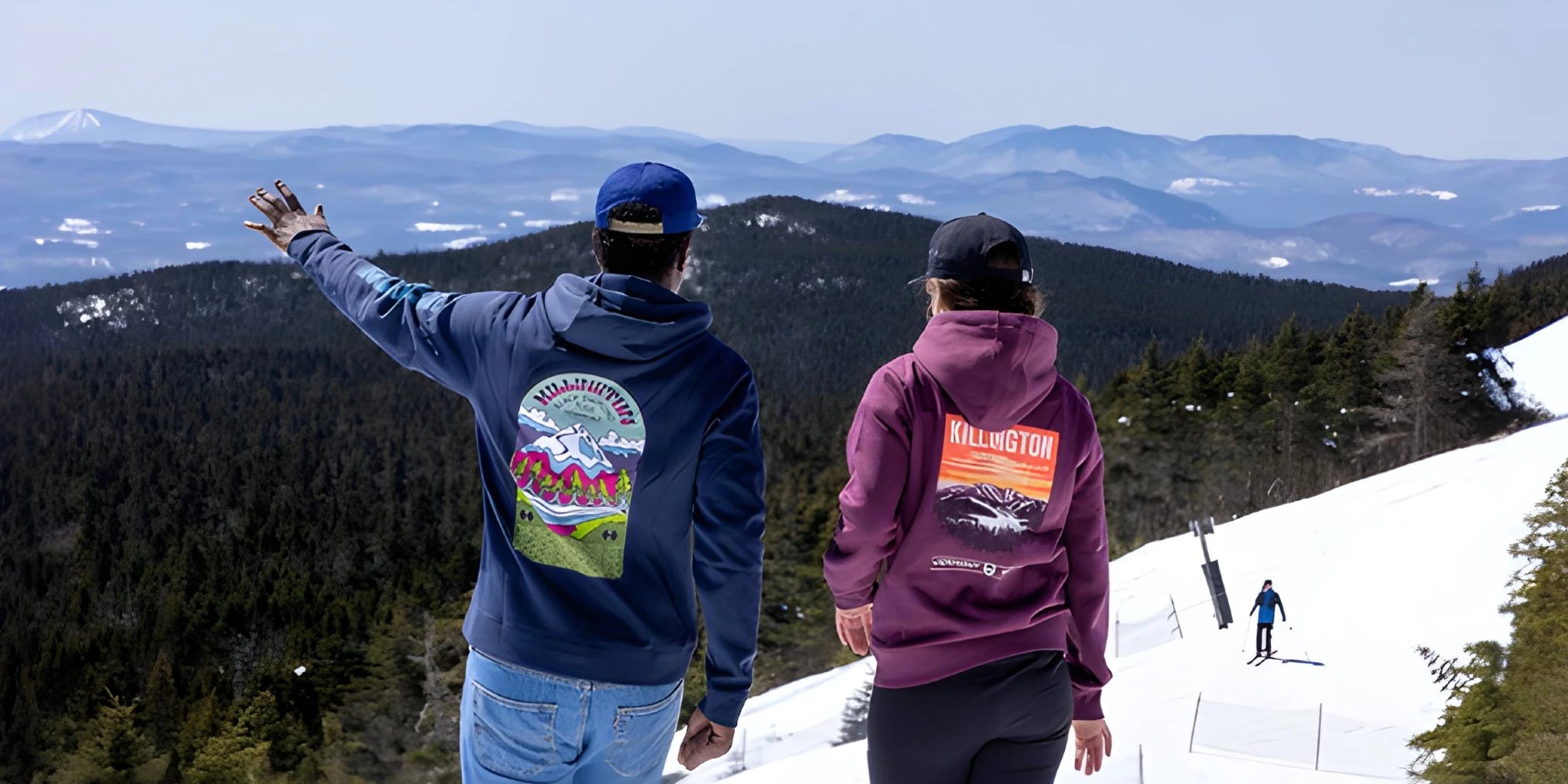 Two people standing on a snowy mountain slope with a scenic view of forested hills and distant blue mountains. The person on the left wears a navy blue hoodie with a large multicolor back graphic and jeans, while the person on the right wears a purple hoodie with a large orange and white back graphic.
